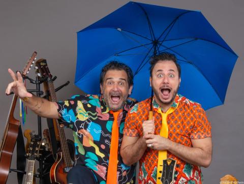 foto de 2 artistas homens com suas bocas abertas, 1 está segurando um guarda-chuva azul aberto e atrás do lado esquerdo tem algumas guitarras