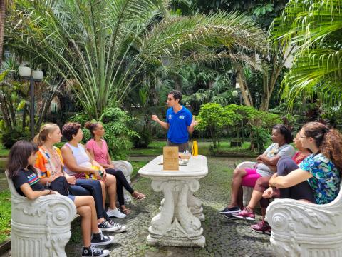 foto de alunos sentados em volta de uma mesa e um educador em pé conversando com eles